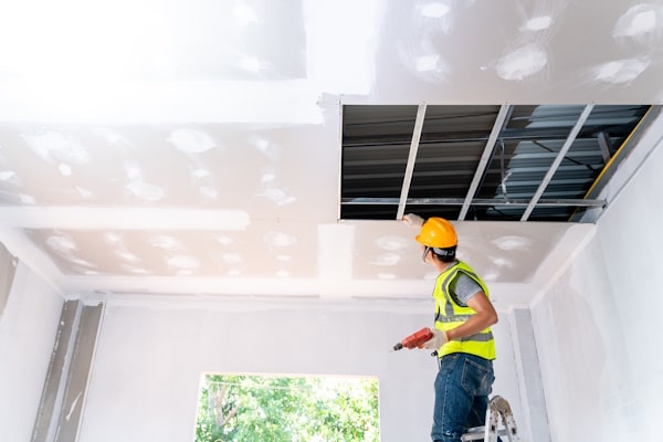 Asian construction worker assemble a suspended ceiling with drywall and fixing the drywall to the ceiling metal frame with screwdriver. Renovation, construction and ceiling installation concept.